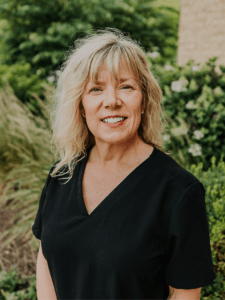 Sarah Melville, front office staff at Evolve Dental Care, smiling in black scrubs with greenery in the background, showcasing a friendly and welcoming demeanor.