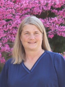 Gina Dunkum, office manager at Evolve Dental Care, smiling in front of vibrant pink flowers, representing her 30 years of experience in dentistry.