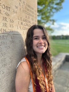 Young woman smiling in front of a stone monument, wearing graduation cords, embodying the supportive and friendly atmosphere at Evolve Dental Care.